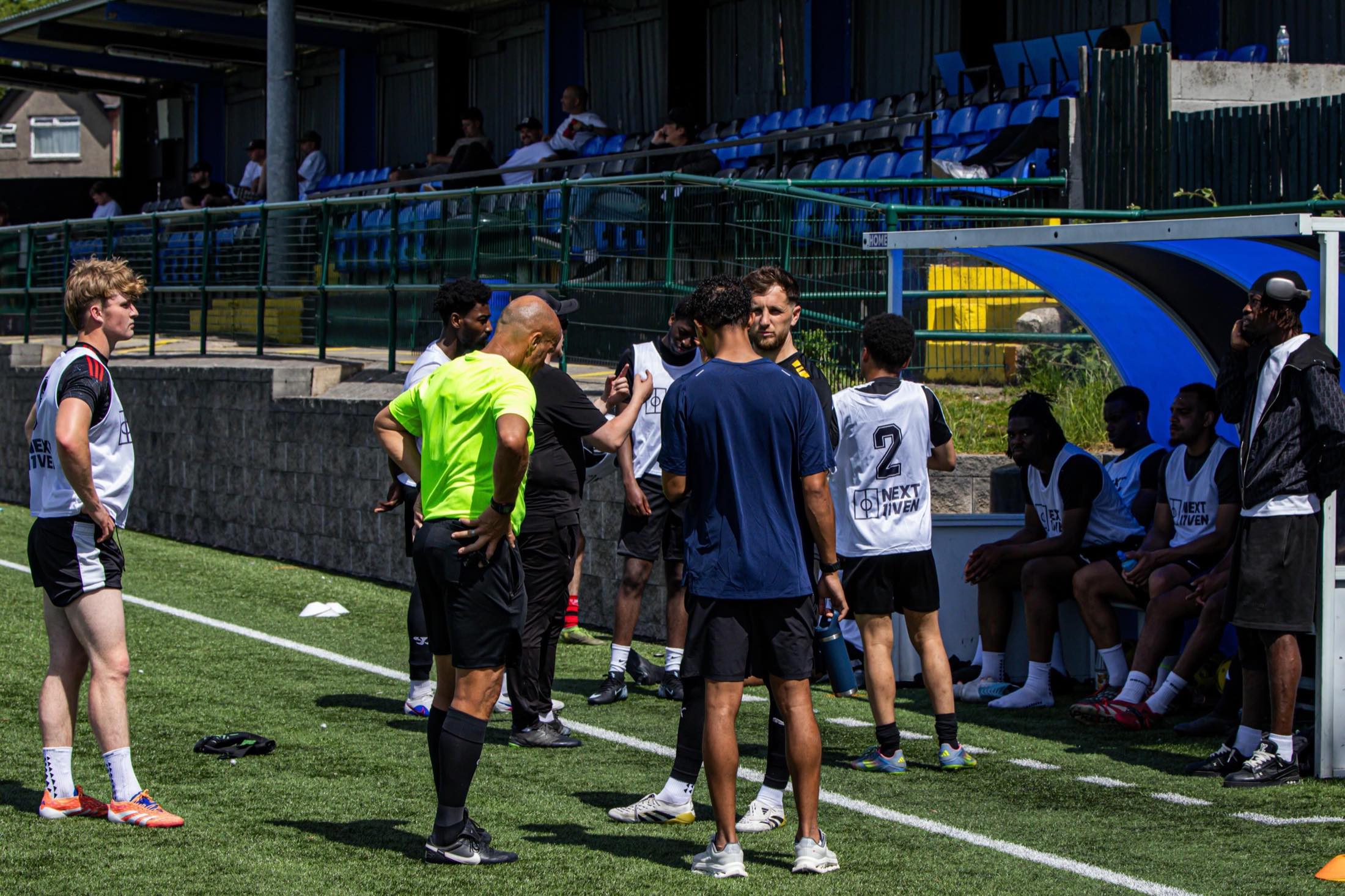 Players in NEXT11VEN bibs at a trial day on a non-league ground, a coach giving a team talk on the touchline.
